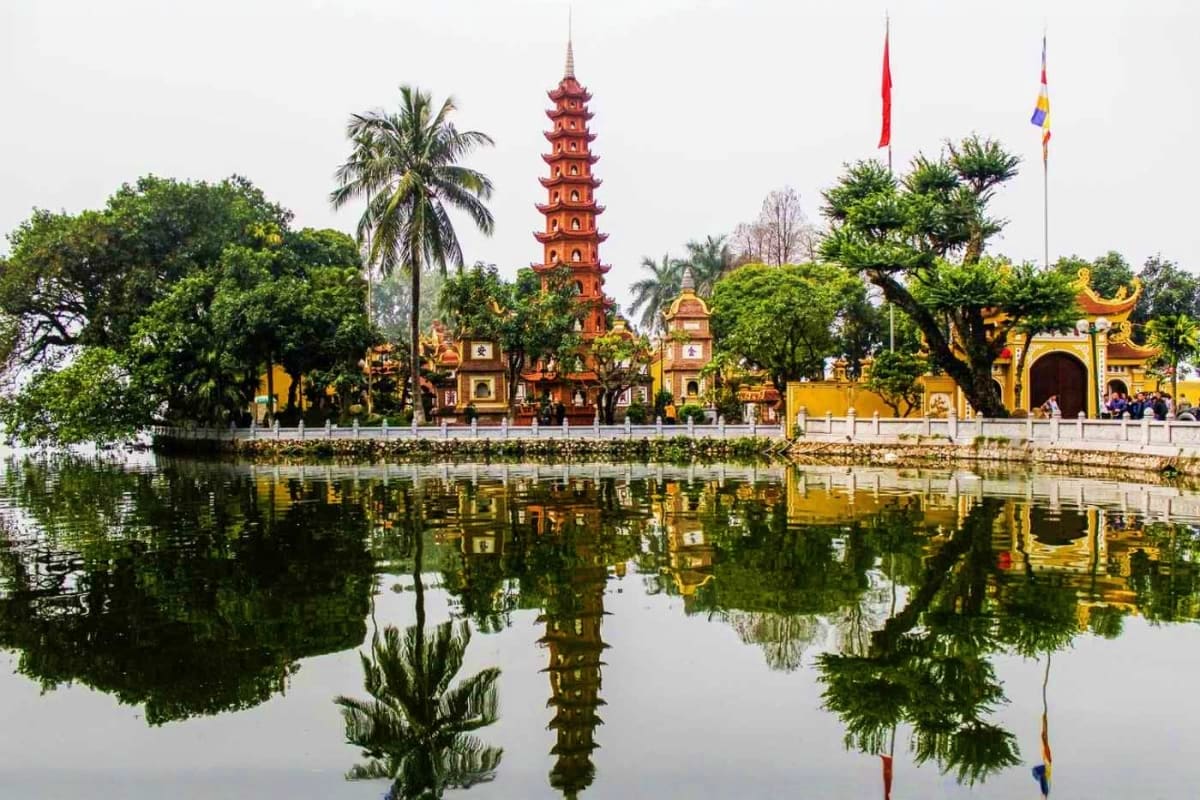 Tran Quoc Pagoda with red brick tower reflected in Hoan Kiem Lake surrounded by yellow temple gates and green trees in Hanoi