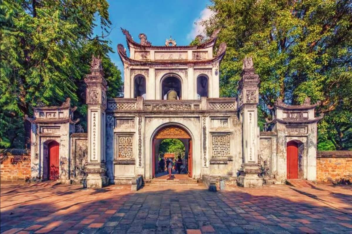 Traditional Vietnamese temple gateway with curved roof, decorative pillars, and red doors framed by green trees in Hanoi