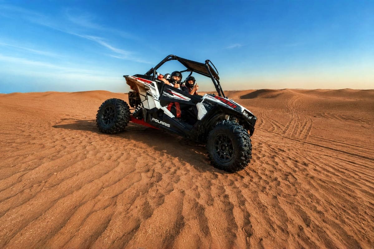 Two people in protective gear driving a red and white Polaris dune buggy across the sandy desert in Dubai under a clear blue 