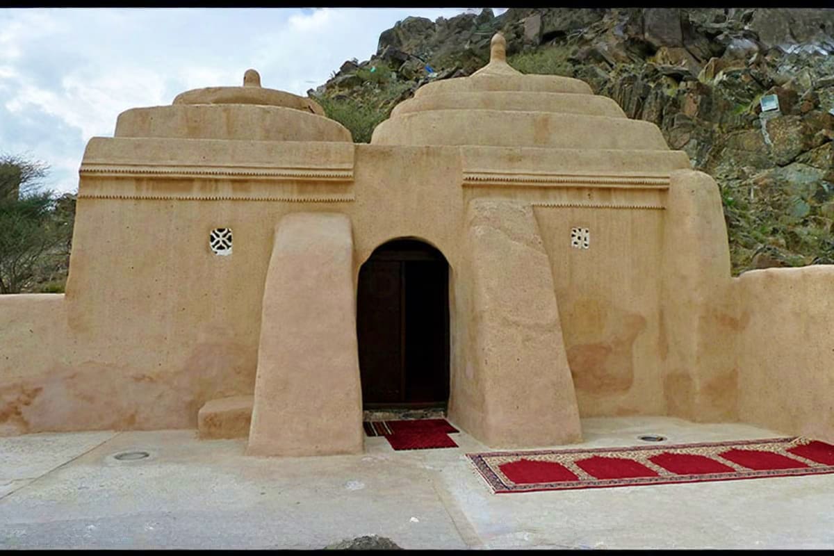 6 Emirates In A Day Tour Ancient clay dome structure with arched entrance and decorative crosses, featuring red carpets and rocky mountain backdrop