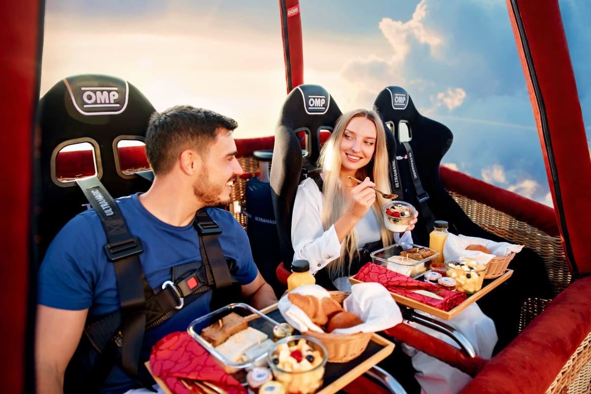 Breakfast in the sky with Balloon flights Couple enjoying breakfast in a hot air balloon basket with food and drinks against a blue sky background