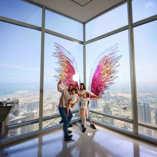 Three visitors posing with pink feathered wing art installation at Burj Khalifa observation deck with aerial cityscape view