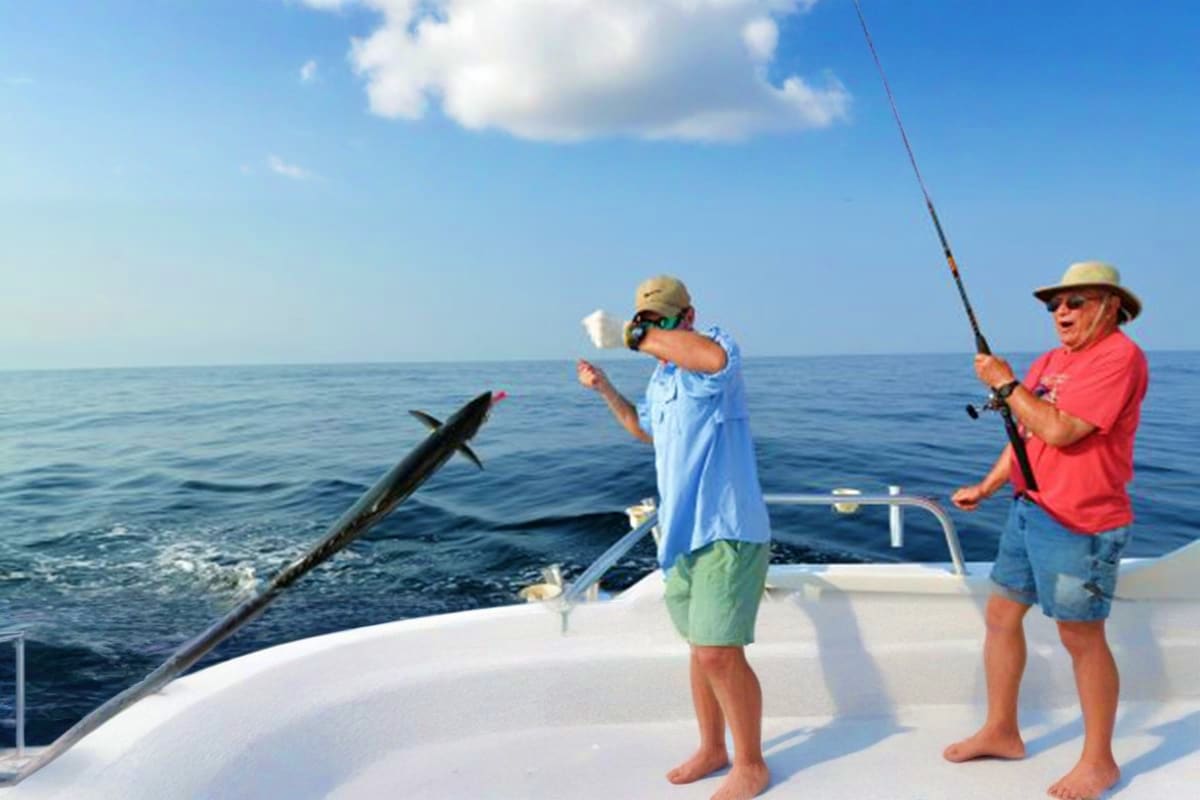 Two men deep sea fishing on a charter boat, one holding a fishing rod with a caught fish jumping out of the water