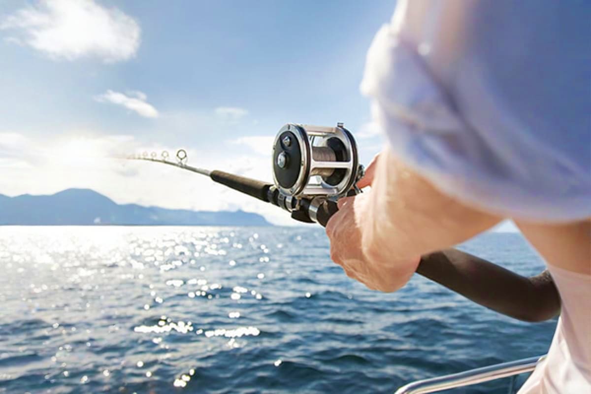 Person holding a fishing rod and reel over ocean water with mountains visible in the distance on a sunny day