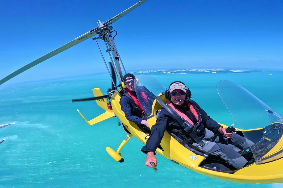 Two passengers in a yellow gyrocopter flying over turquoise ocean waters with coral reefs visible below