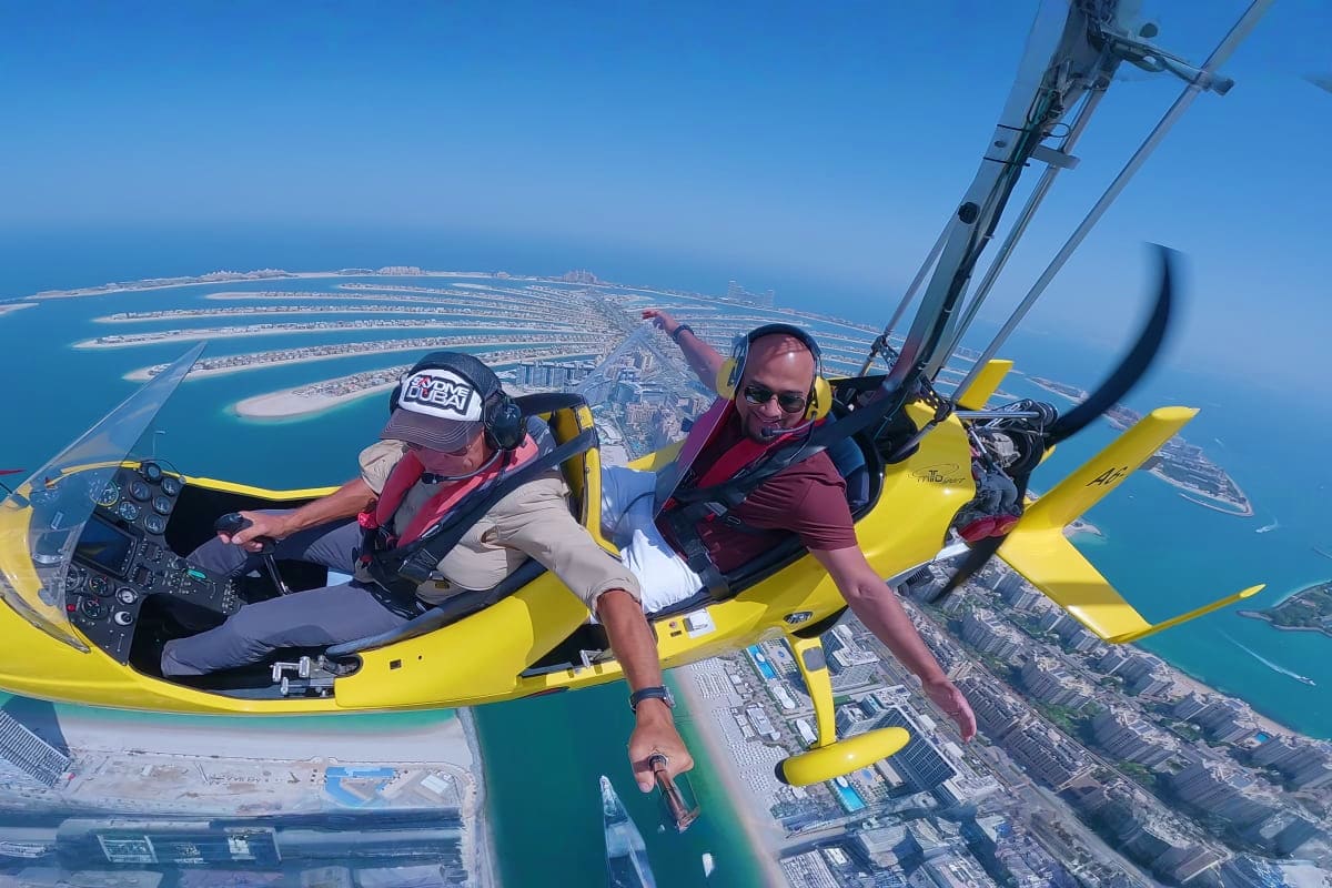 Two people in yellow gyrocopter flying above Dubai's Palm Jumeirah island and turquoise coastline waters
