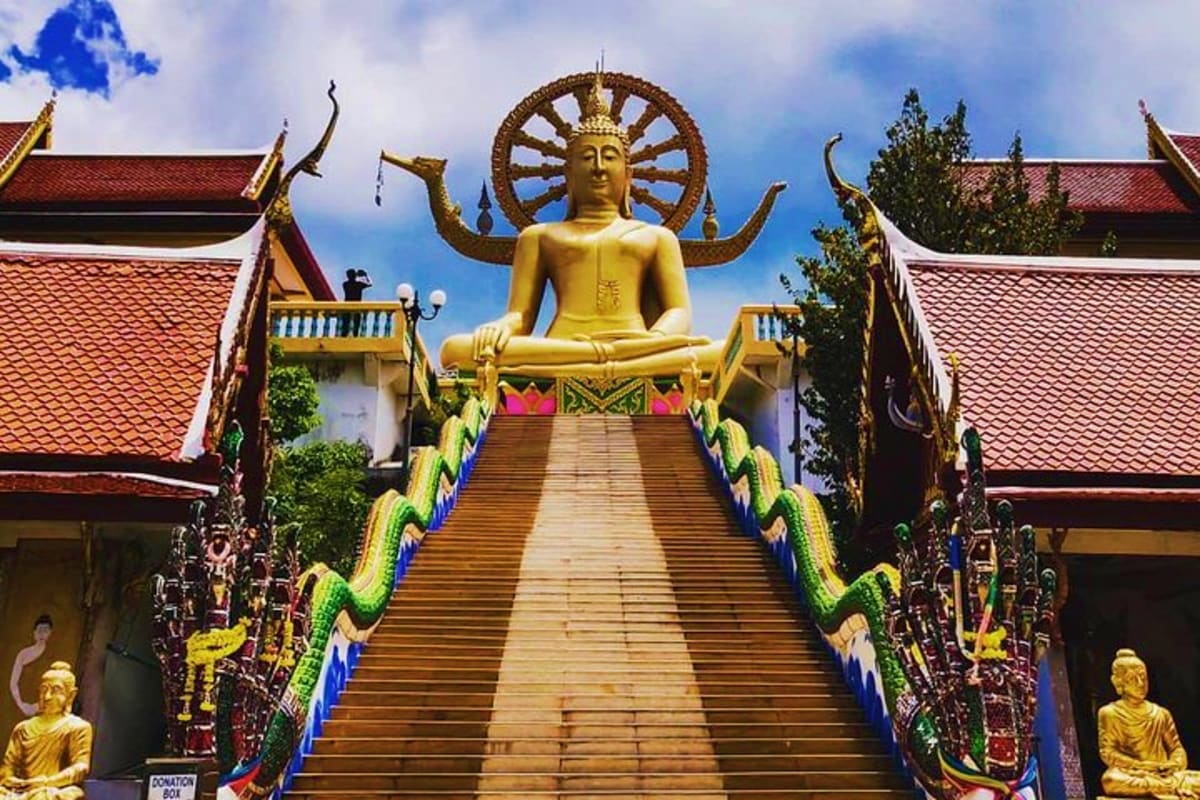 Giant golden Buddha statue with decorative wheel halo at top of temple stairs flanked by green dragon railings and traditiona