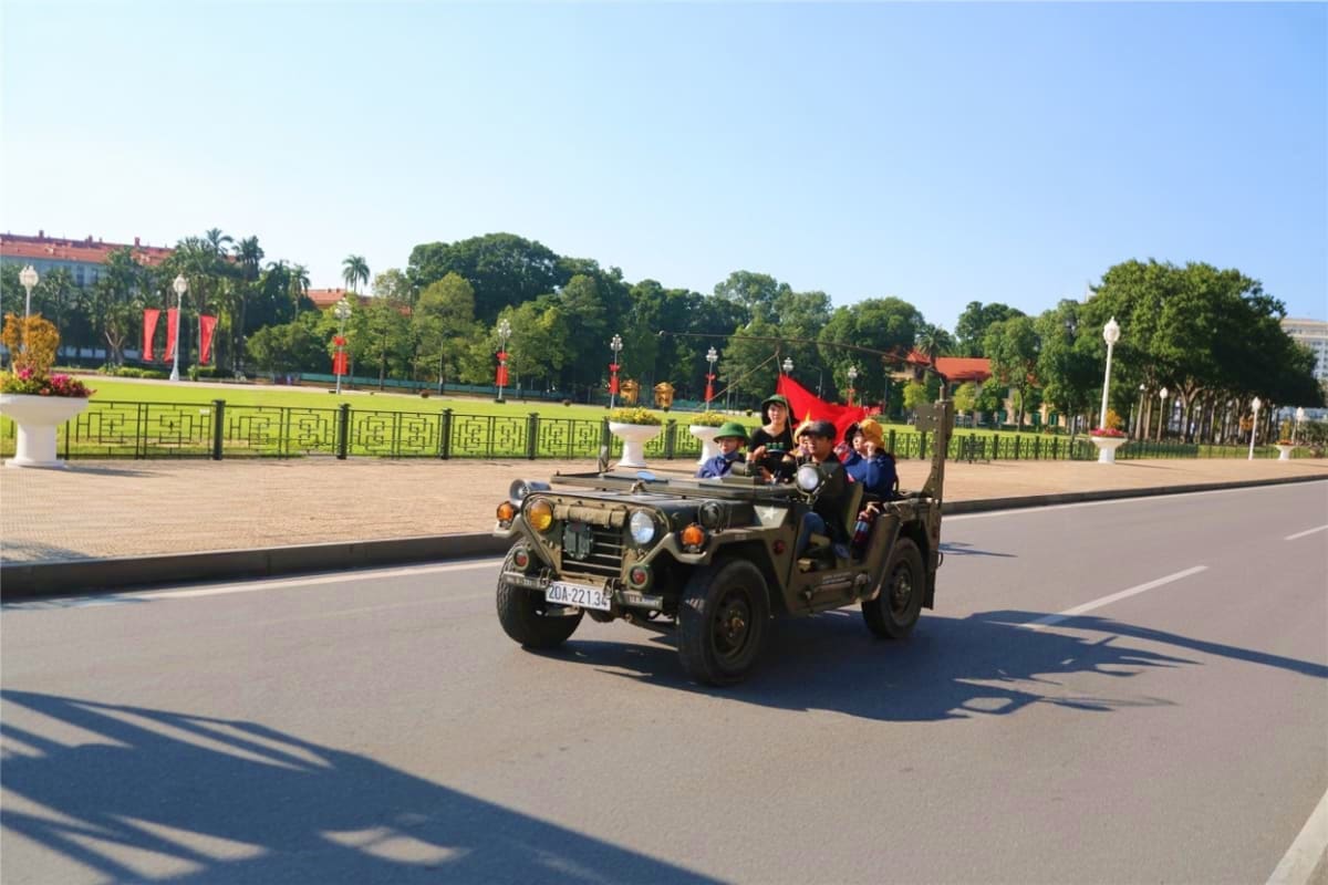 Vintage military jeep carrying tourists with red flag driving through Ba Dinh Square in Hanoi with ornate green gates and tre