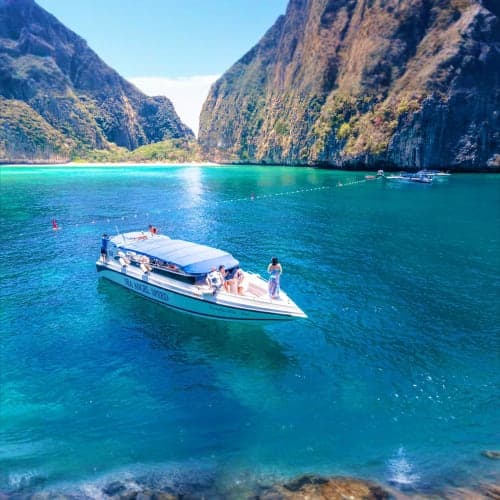 Speedboat anchored in turquoise waters beneath tall limestone cliffs at Hong Island, Thailand, with tourists on board and in 