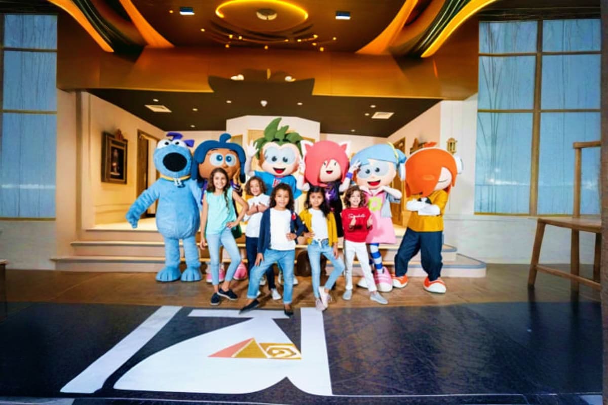 KidZania Abu Dhabi Group of children posing with colorful cartoon character mascots in a modern indoor theme park lobby with orange and black ce