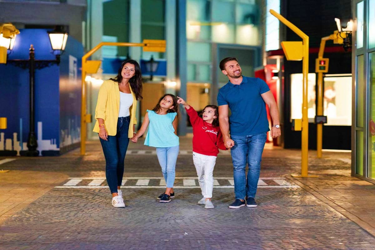 KidZania Abu Dhabi Family of four walking through illuminated urban street at night with yellow architectural structures and storefronts in back
