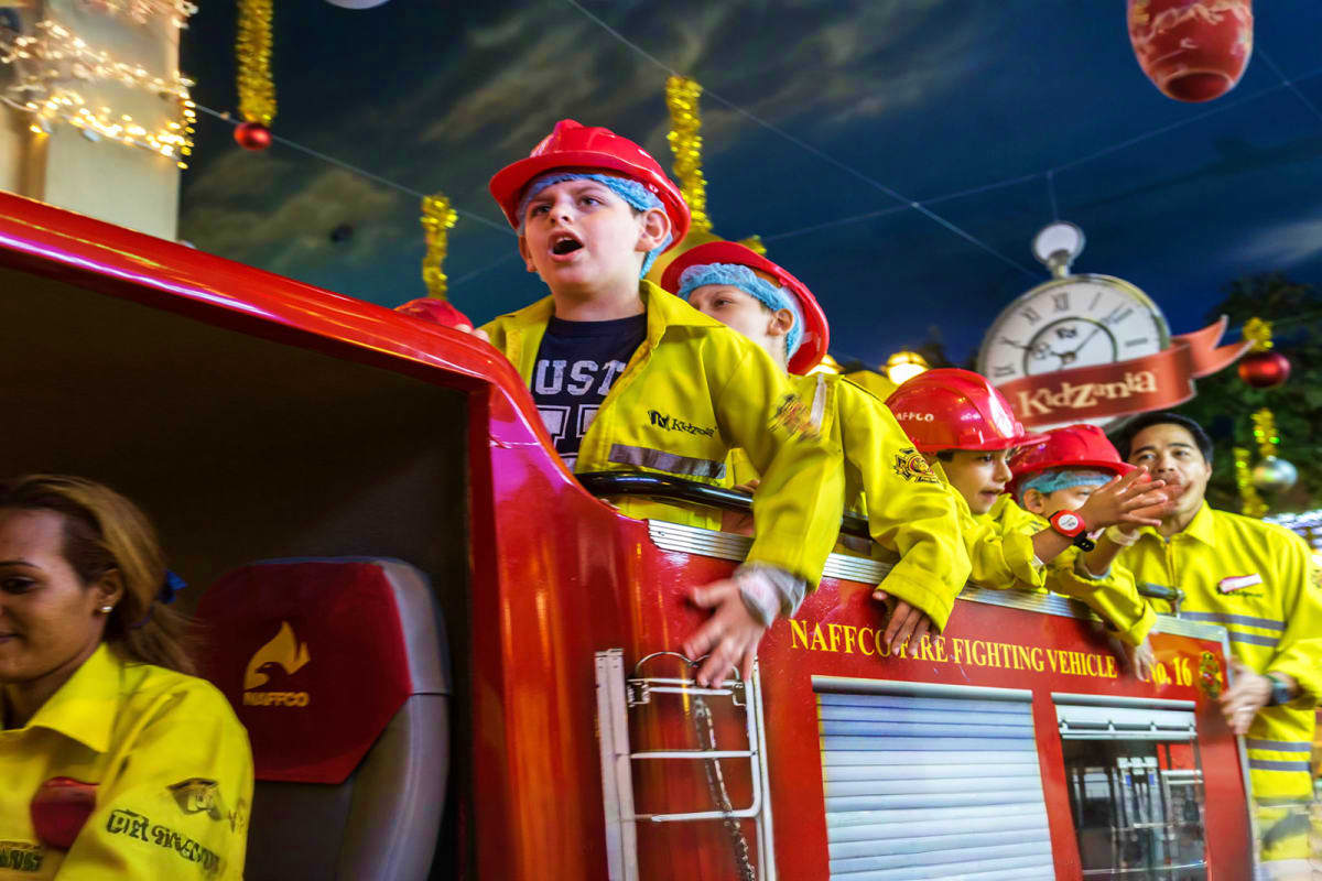 KidZania Abu Dhabi Children in firefighter uniforms and red helmets riding on a toy fire truck attraction at KidZania Abu Dhabi indoor theme par