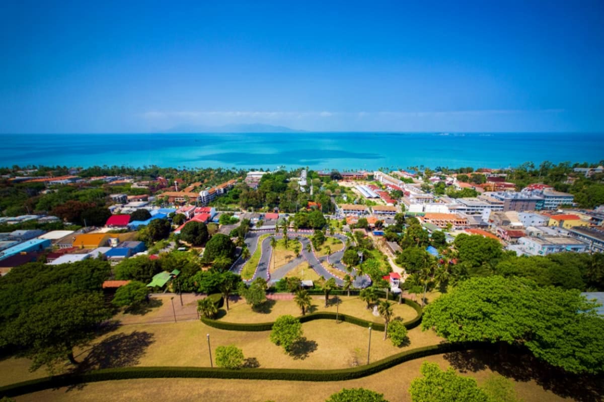 Koh Samui Half Day City Tour Aerial view of Koh Samui coastal town with turquoise ocean, palm trees, colorful buildings, and green spaces along the shorel
