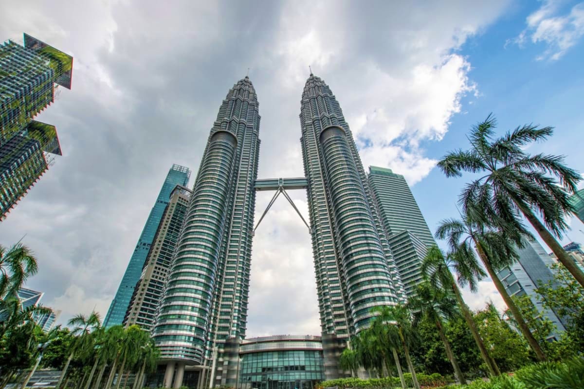 Petronas Twin Towers in Kuala Lumpur with connecting skybridge, surrounded by palm trees and modern buildings under cloudy sk