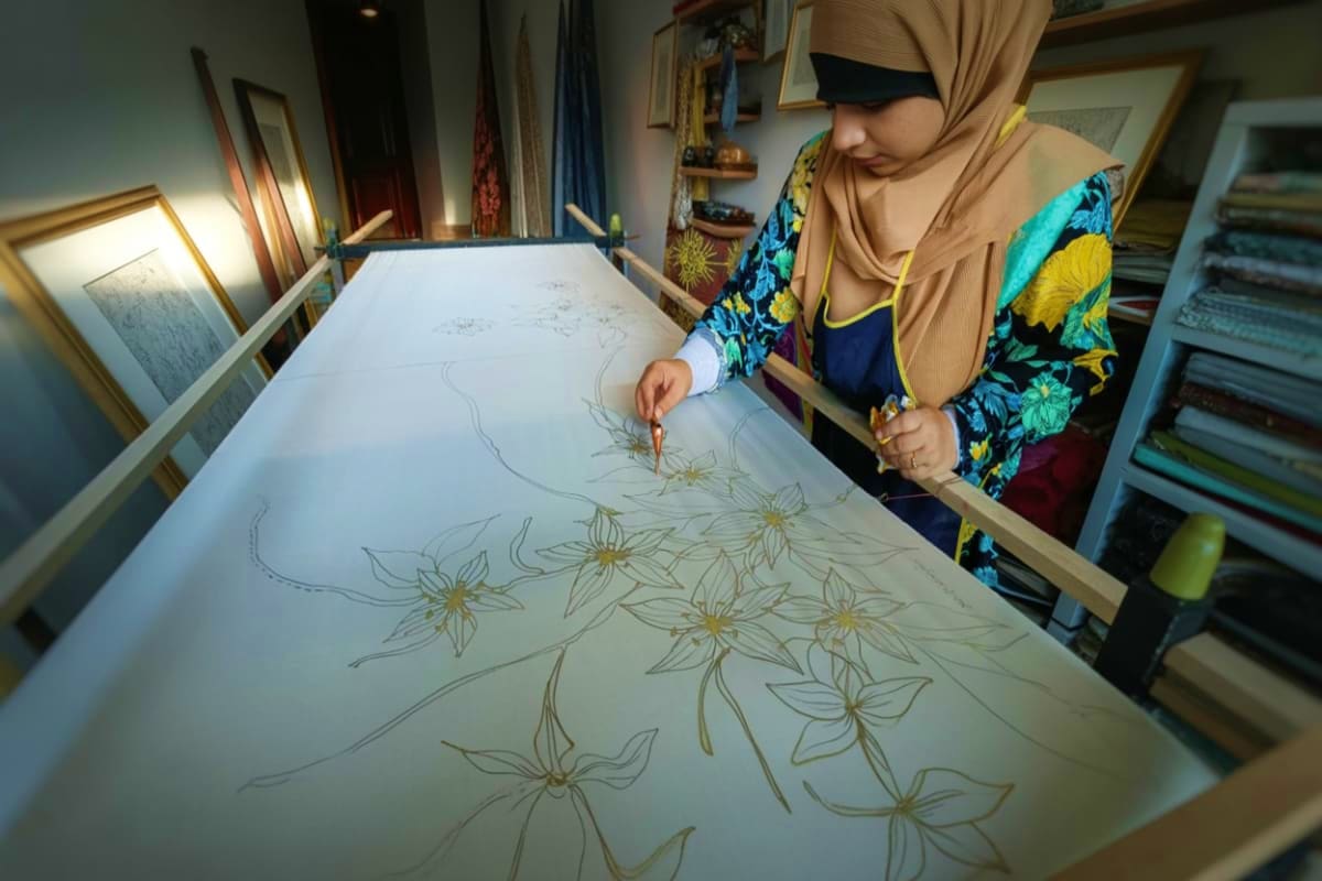 Woman drawing floral patterns on large white paper at a design studio workstation with art supplies and framed artwork visibl