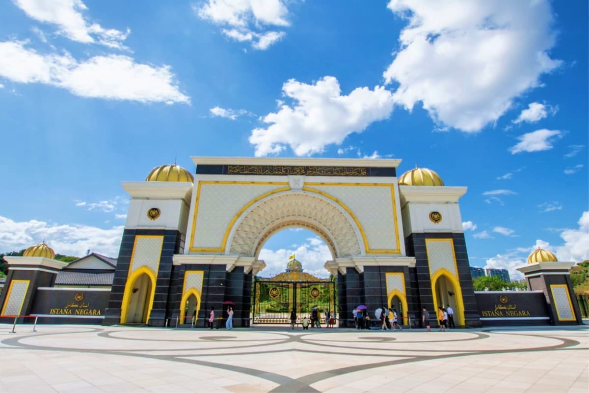 Istana Negara palace gates with golden domes, white arches, and black and gold accents under blue sky with white clouds in Ku