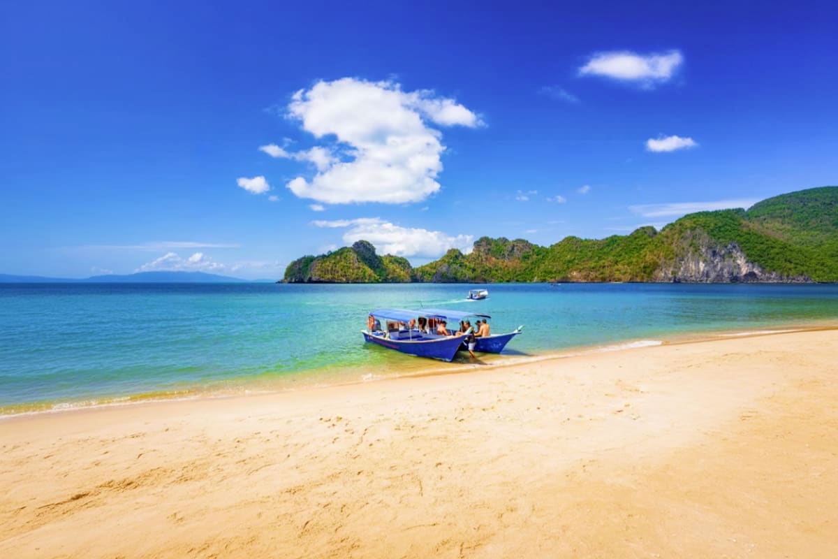 Blue fishing boat anchored on sandy beach with turquoise water and forested islands under blue sky in Langkawi