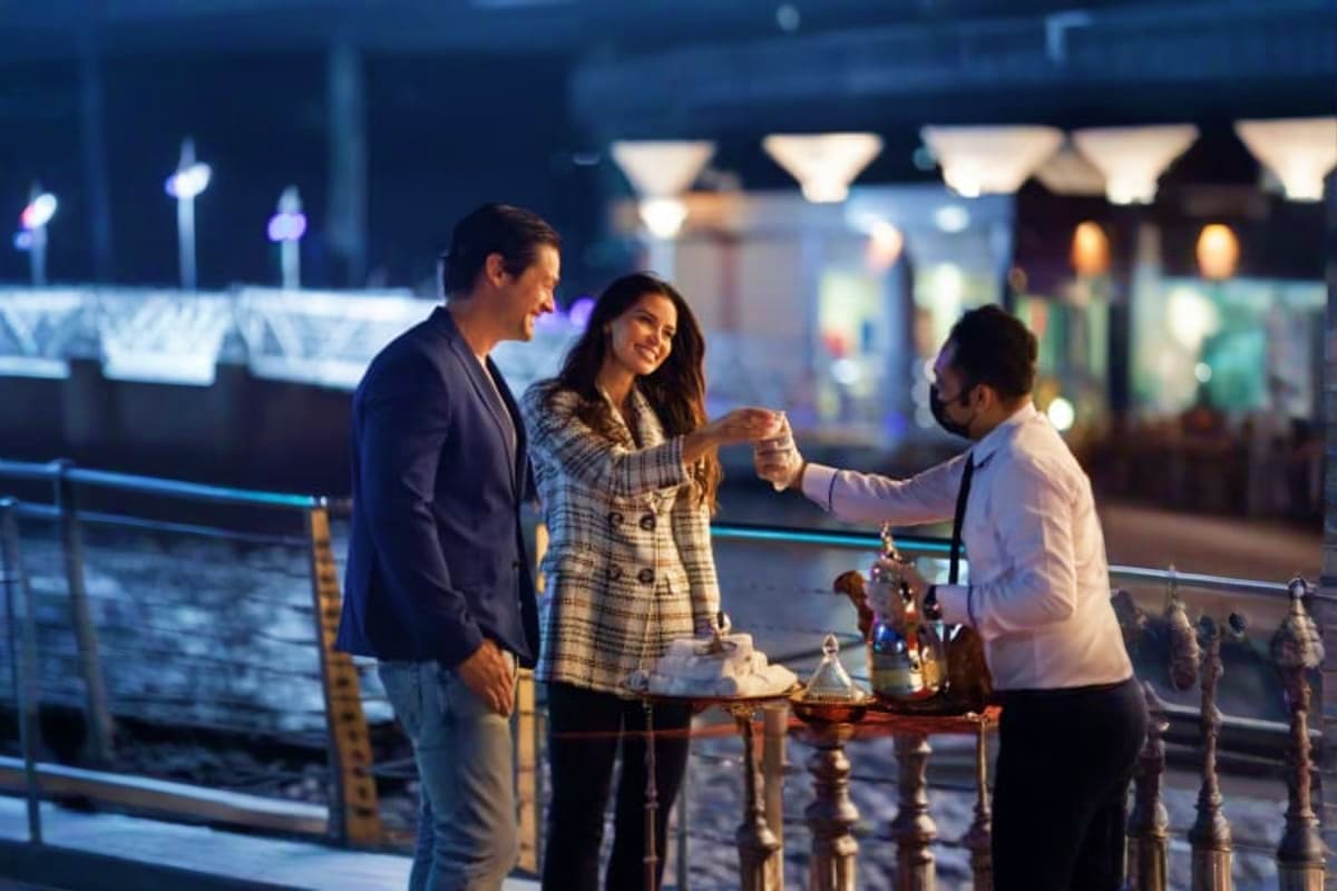 Luxury Dubai Canal Cruise Three people enjoying drinks on an illuminated Dubai waterfront promenade at night with city lights and water views in backgr