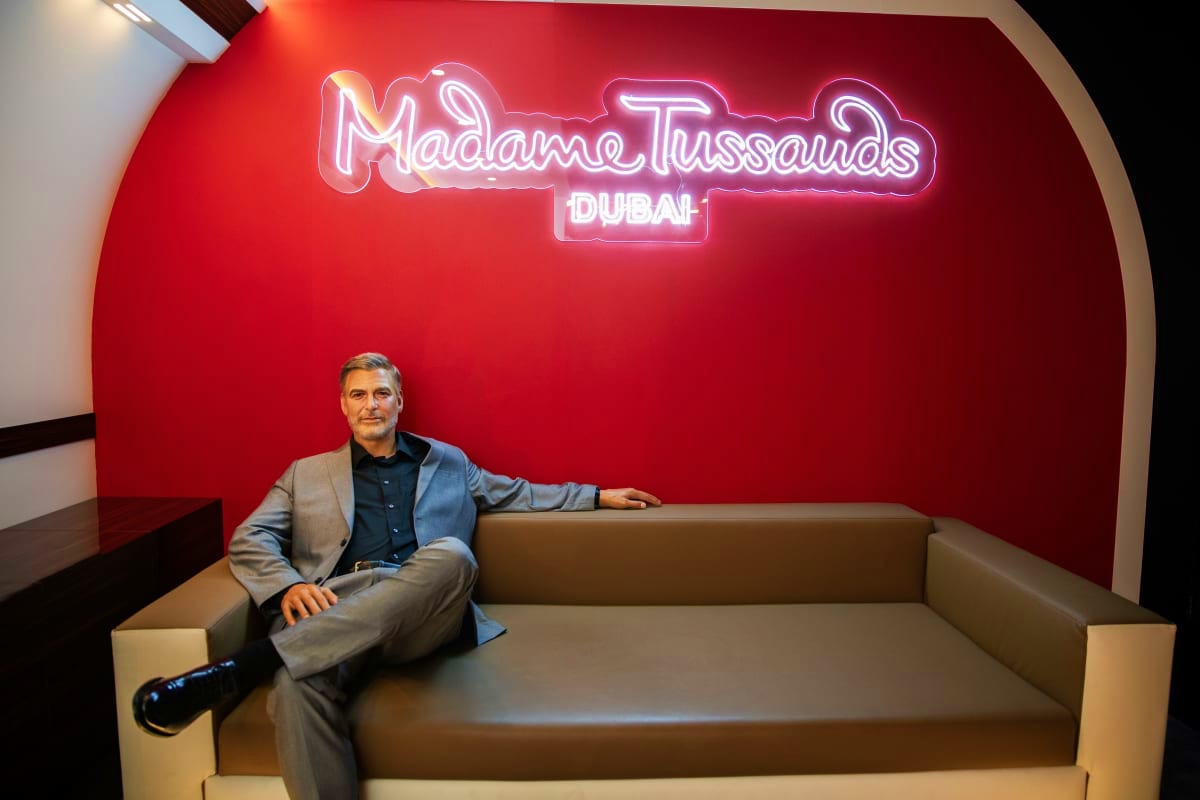 Wax figure of a man in business attire sitting on a beige couch in front of a red wall with Madame Tussauds Dubai neon sign