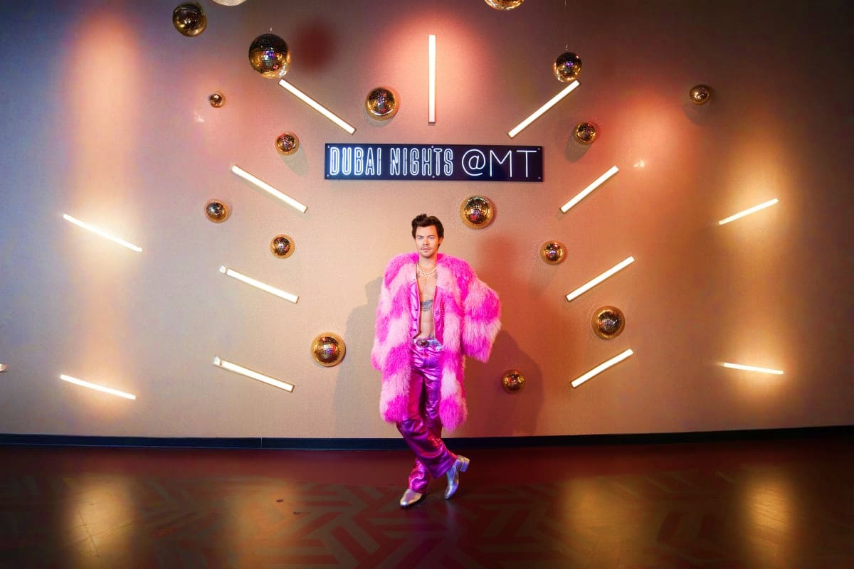 Man in bright pink fur coat and purple pants posing under neon Dubai Nights sign with gold spheres at Madame Tussauds Dubai w
