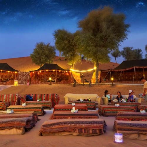 Bedouin desert camp at night with traditional seating, illuminated tents, and guests gathered around a fire under acacia tree