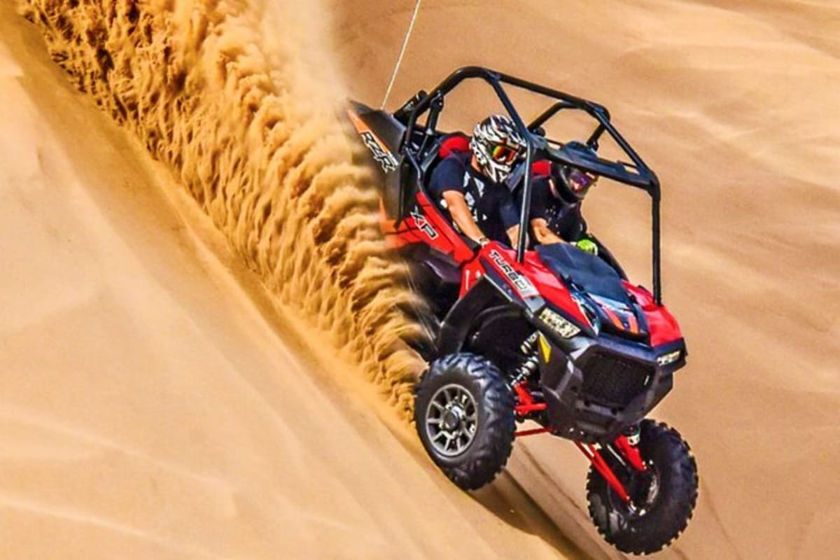 Red and black dune buggy with two riders driving up a steep sandy dune in desert terrain