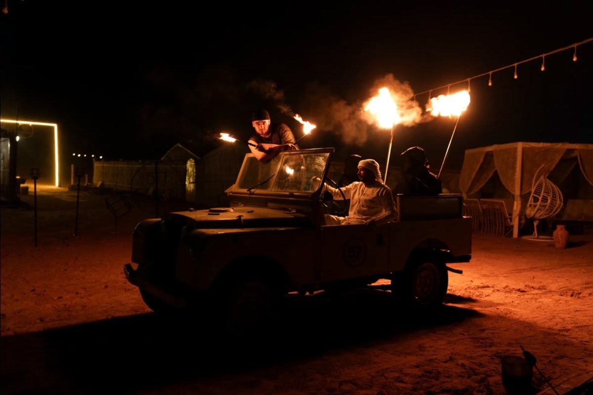 Nighttime desert safari camp with fire performers juggling flaming torches, illuminated by overhead lights and a vehicle in t