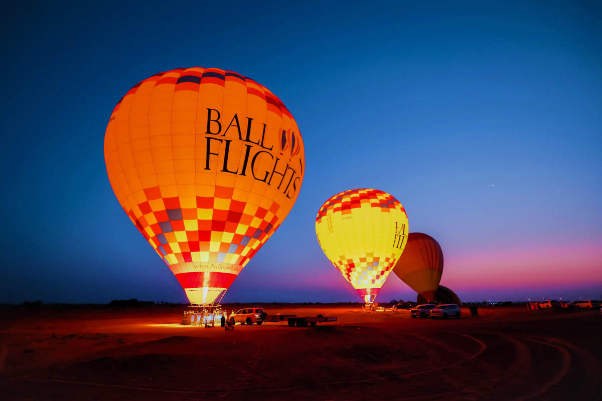 Illuminated hot air balloons on desert ground at dusk with Ball Flights branding visible on envelopes