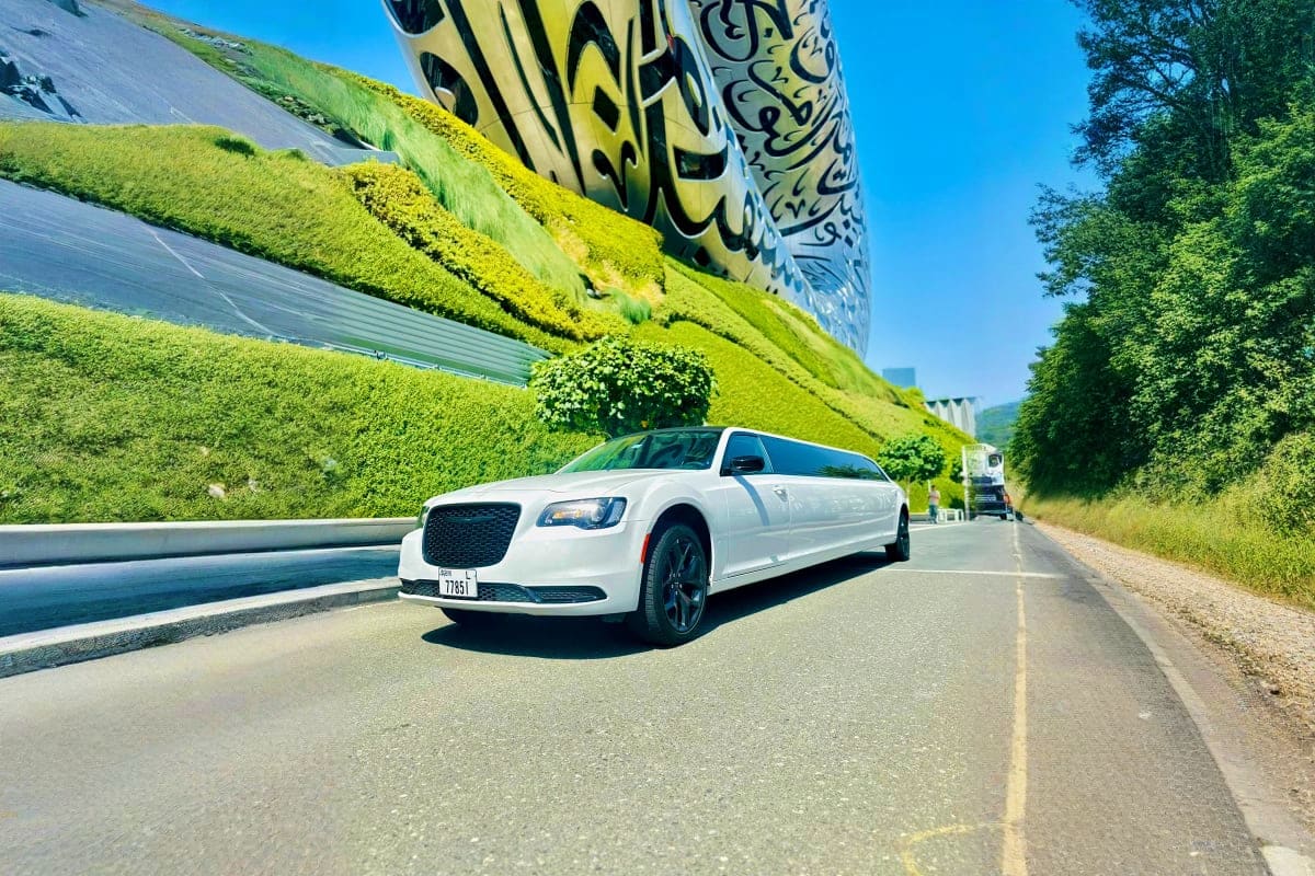 White luxury limousine driving on road past large graffiti-covered cylindrical structure and green hillside under clear blue