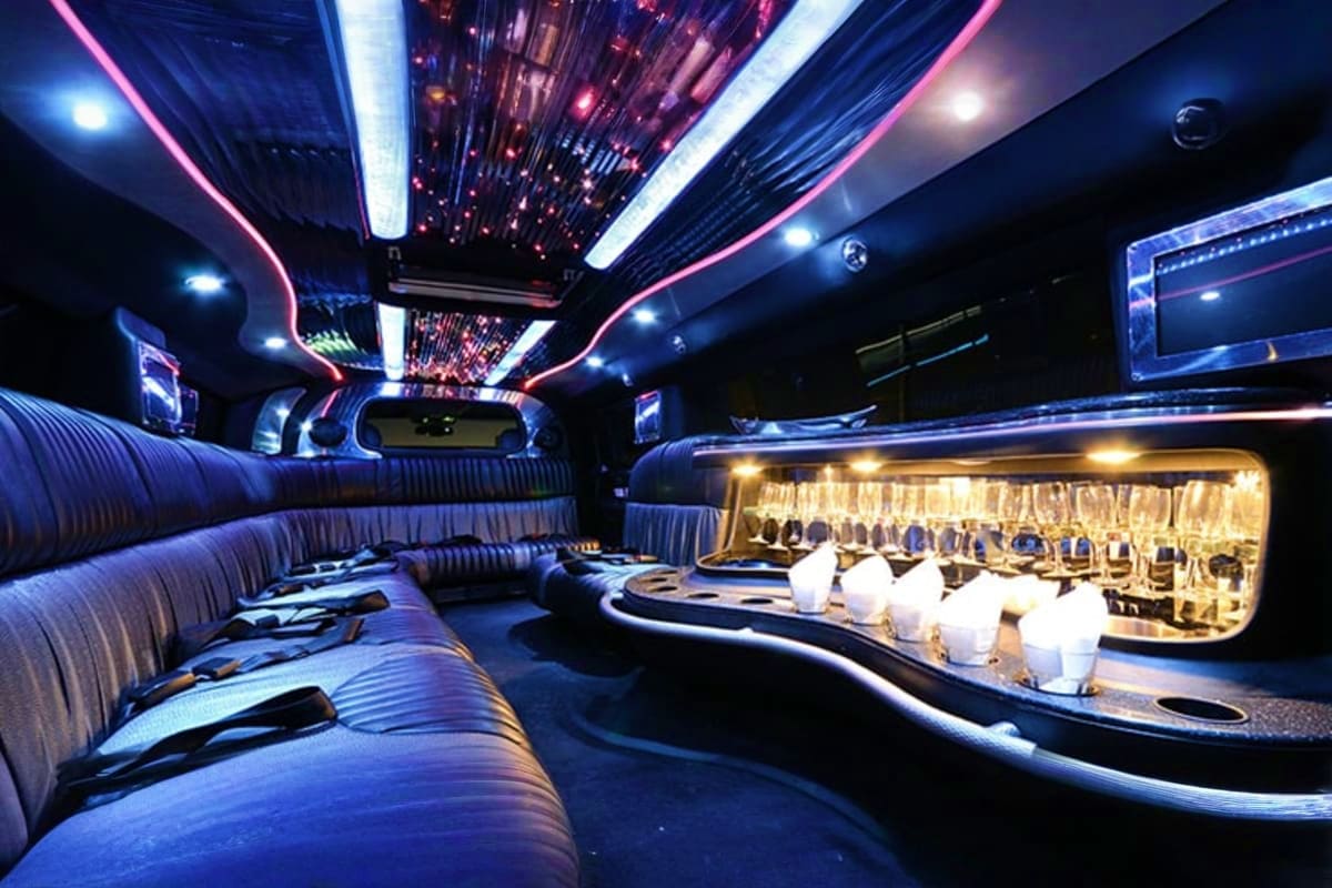 Luxurious limousine interior with neon pink and blue LED lighting, striped seats, bar counter with glasses, and entertainment