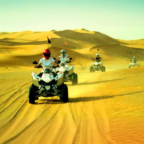 Group of quad bikers riding across golden sand dunes in Ras Al Khaimah desert with flags mounted on vehicles