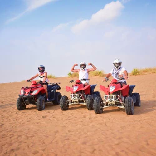 Quad Bike In Ras Al Khaimah Three riders on red quad bikes lined up in desert sand with dunes and sky backdrop in Ras Al Khaimah