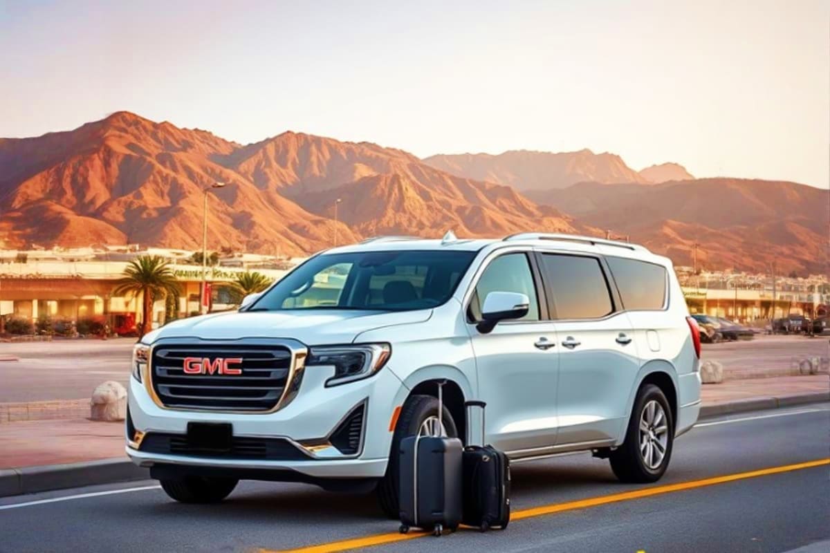 White GMC minivan with luggage parked on road with desert mountains in background, representing airport transfer service