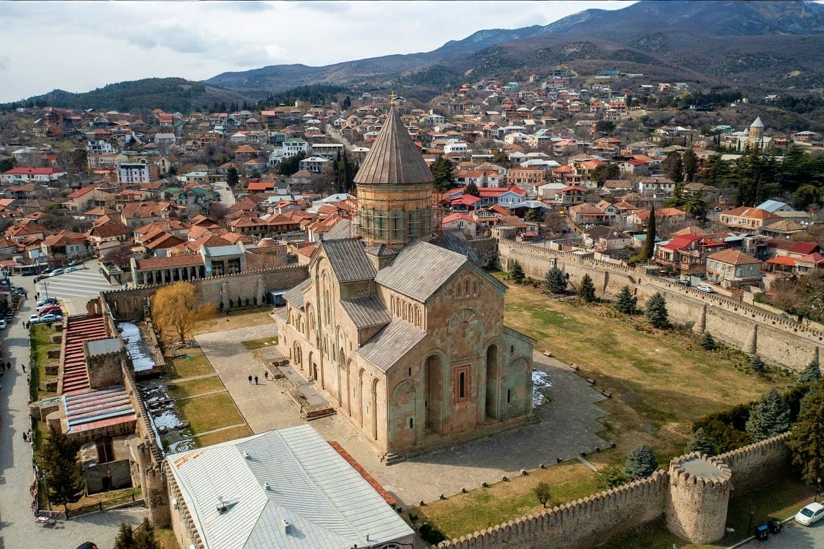 Aerial view of Svetitskhovlsi Cathedral in Mtskheta with stone fortress walls, traditional red-roofed houses, and mountains i