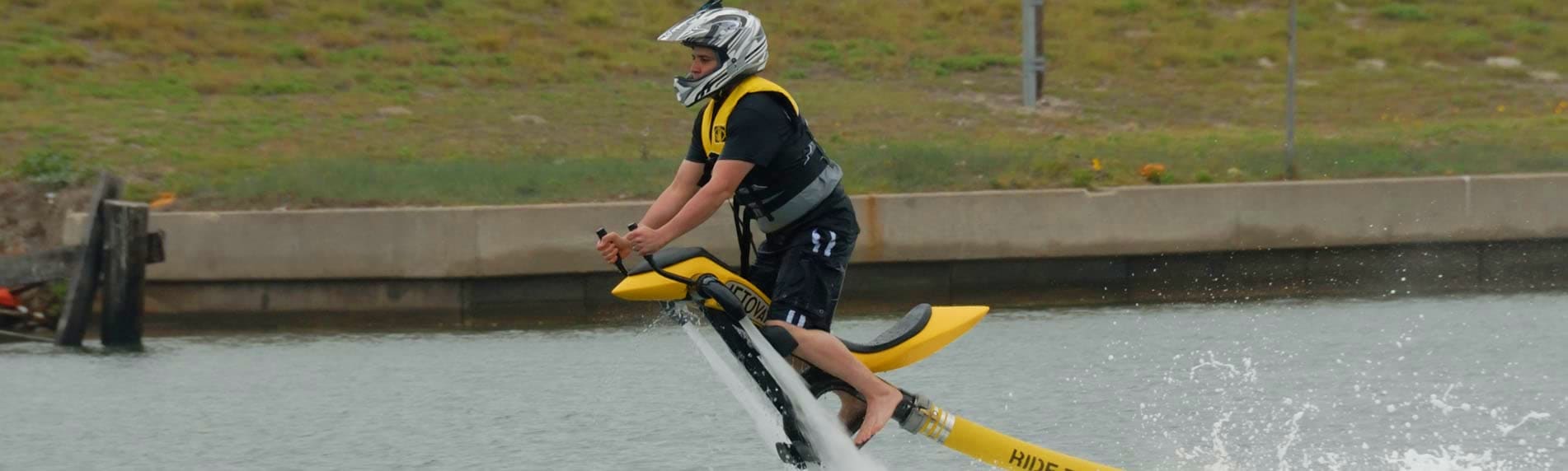 Jetovator Tour Dubai Rider performing jetovator water sport maneuver on yellow jet ski in calm water with concrete embankment and grass field in b