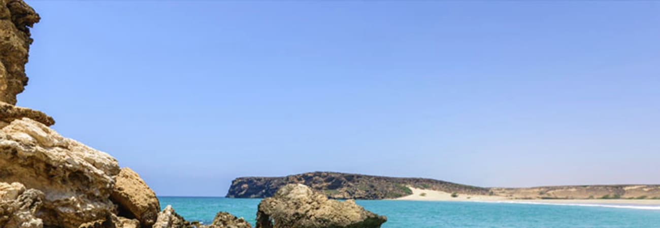 Rocky limestone coastline with turquoise waters and sandy beach on Damaniyat Island under clear blue sky