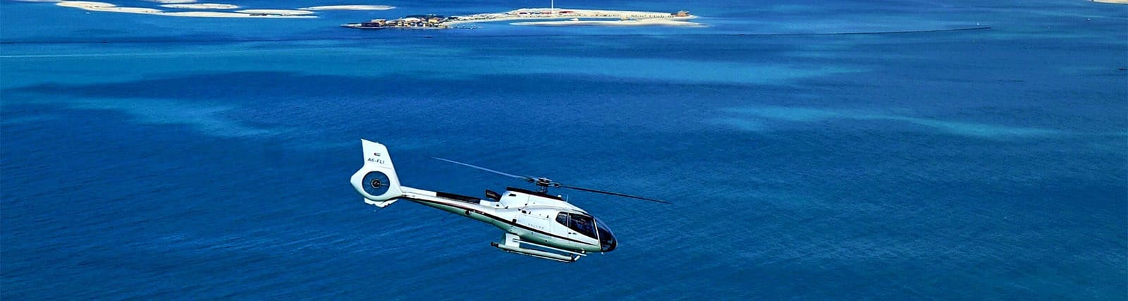 Private Helicopter Tour in Dubai White helicopter flying over turquoise ocean waters with sandy island visible in background