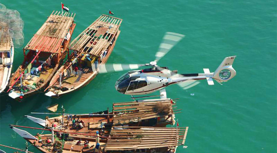 Private Helicopter Tour in Dubai Aerial view of traditional wooden dhow boats and a white helicopter over turquoise water in Dubai