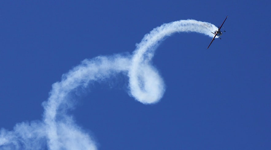 Aerobatic aircraft performing a looping maneuver with white smoke trail against clear blue sky