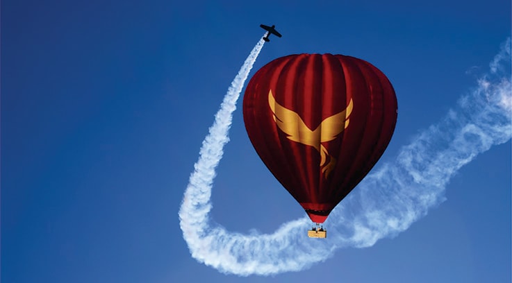 Sunrise Hot Air Balloon Ras Al Khaimah Red and gold hot air balloon floating in blue sky with aerobatic aircraft performing curved smoke trail maneuver beside it