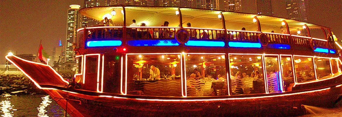 Dhow Cruise Dinner Marina Illuminated traditional red dhow cruise ship at night on Marina waters with passengers dining inside lit cabin and Dubai skyl