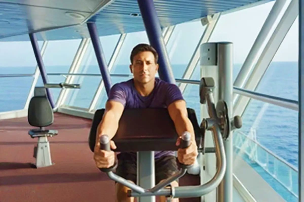 Man exercising on stationary bike in ship's fitness center with ocean view through windows