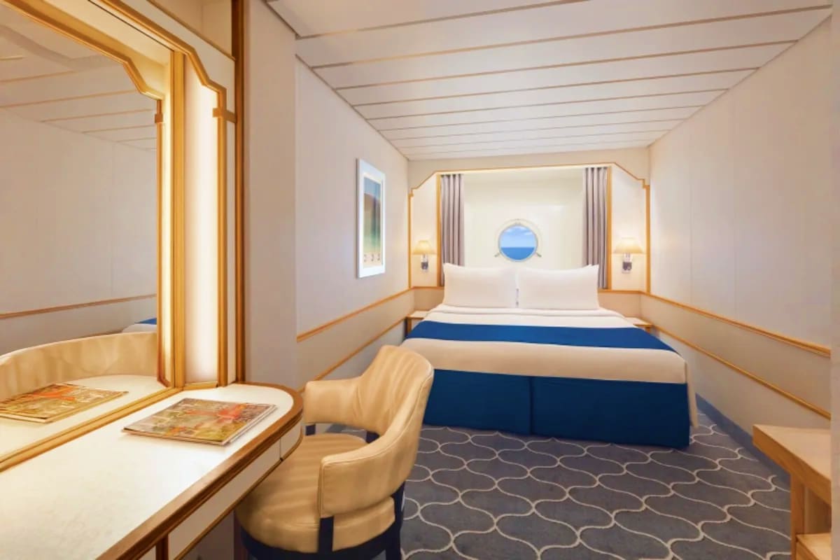 Modern cruise ship cabin with queen bed, white linens, blue bedding, porthole window with ocean view, desk with chair, and go