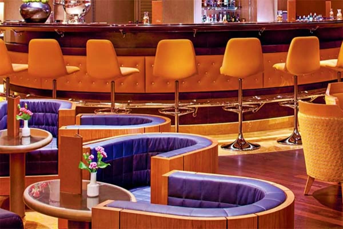 Luxurious cruise ship bar with orange upholstered bar stools, dark wood counter, booth seating with purple cushions, and deco