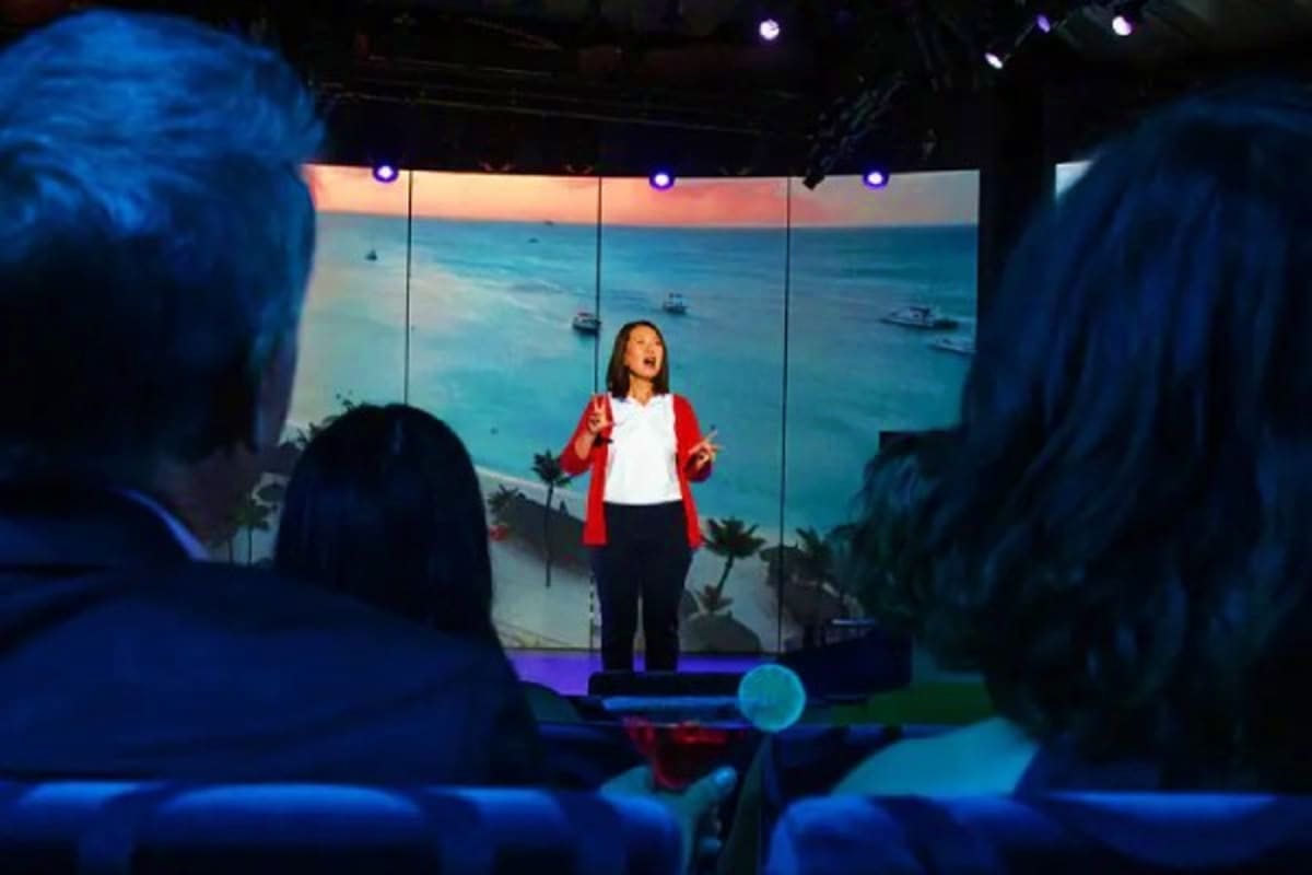 Woman speaking on stage in red jacket with large screen displaying tropical ocean sunset and boats behind her during cruise p