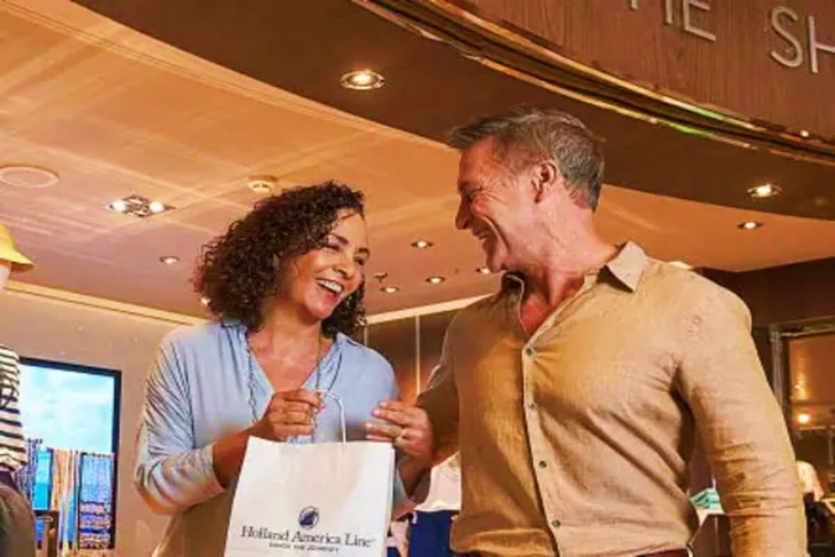 Two people smiling while exchanging a Holland America Line shopping bag in an elegant ship interior with curved ceiling and r