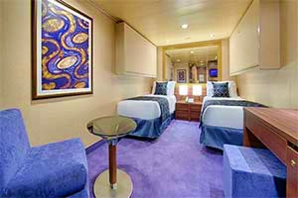 Luxury cruise ship cabin with two twin beds, blue armchair, round side table, wood dresser, and abstract blue and gold artwor