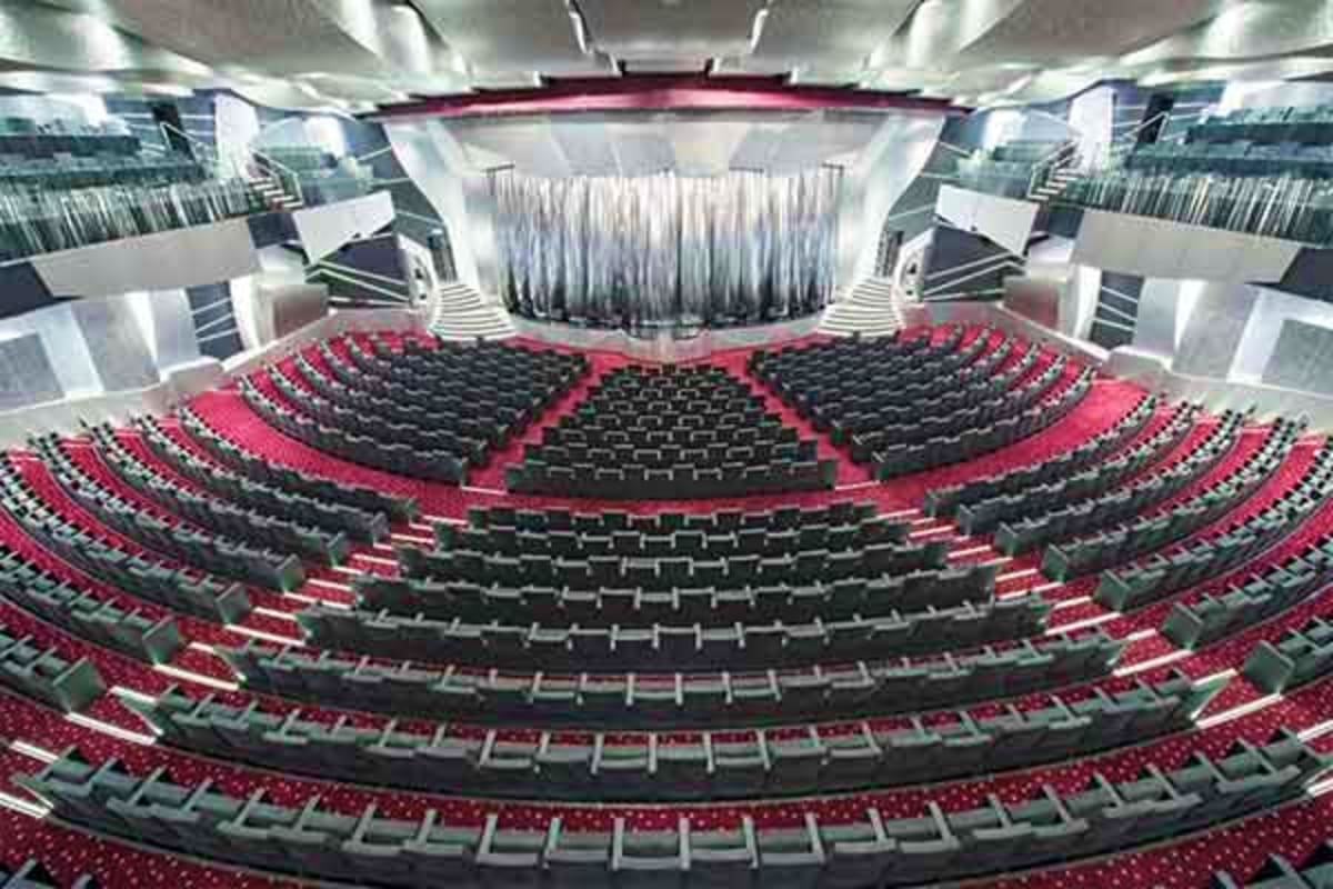 Modern theater interior with rows of black seats, red carpeting, and white curtains on stage under bright lighting