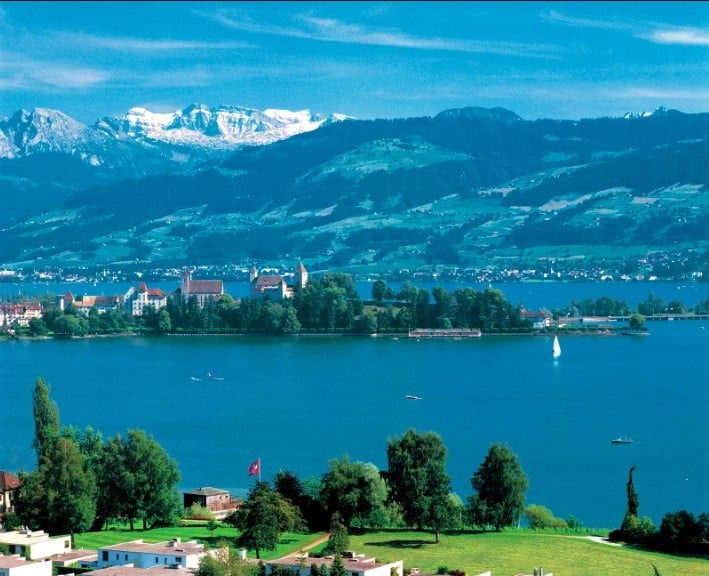 Switzerland Swiss Alpine landscape with blue lake, lakeside town, snow-capped mountains, sailboat, and green meadows in foreground