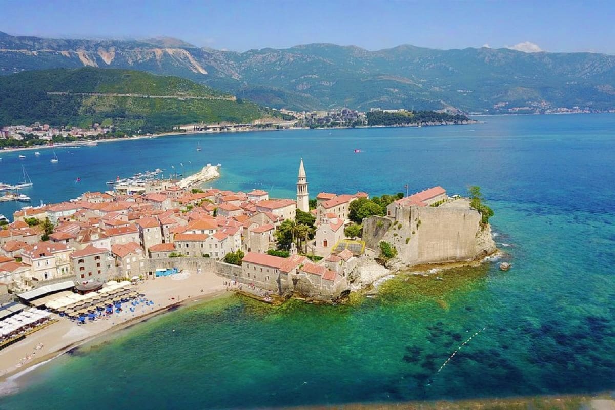 Aerial view of Budva Old Town peninsula with red-roofed stone buildings, church bell tower, turquoise Adriatic Sea, beach, an